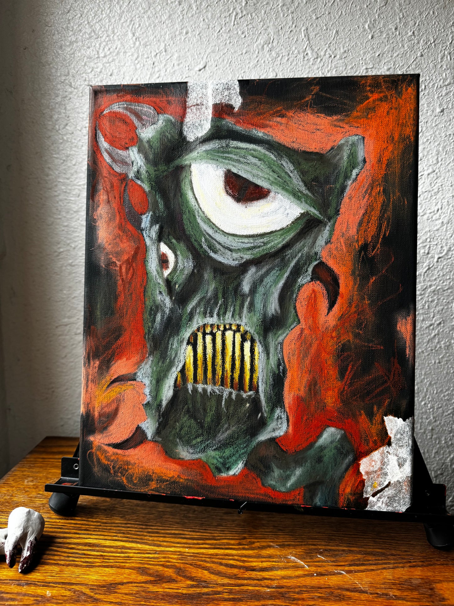 Photo of art piece on an easel, green fantastical monster with horns, yellow teeth, and a very large eye. bckground is black and orange, almost entirely oil pastel.