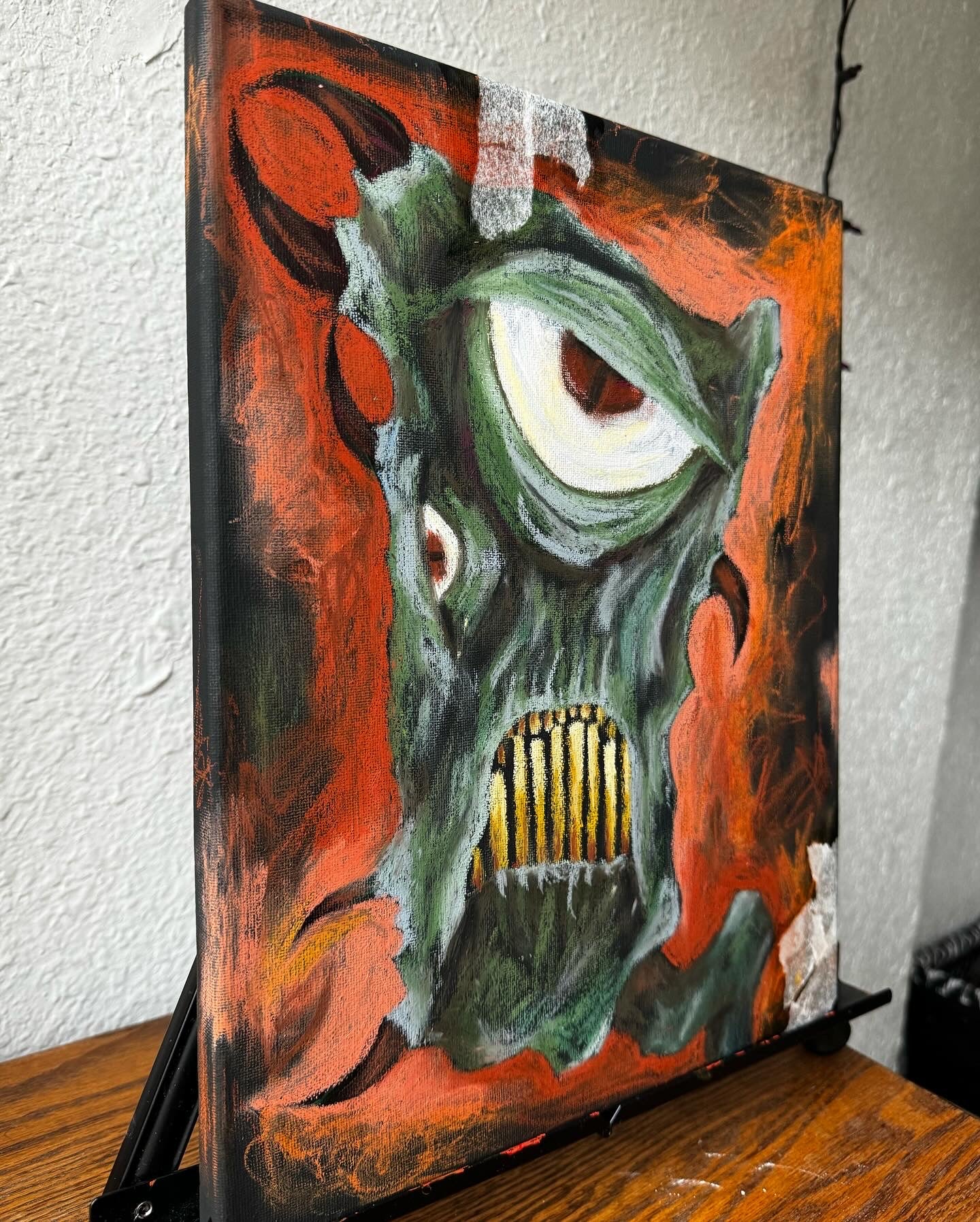 Side angle photo of art piece on an easel, green fantastical monster with horns, yellow teeth, and a very large eye. bckground is black and orange, almost entirely oil pastel.