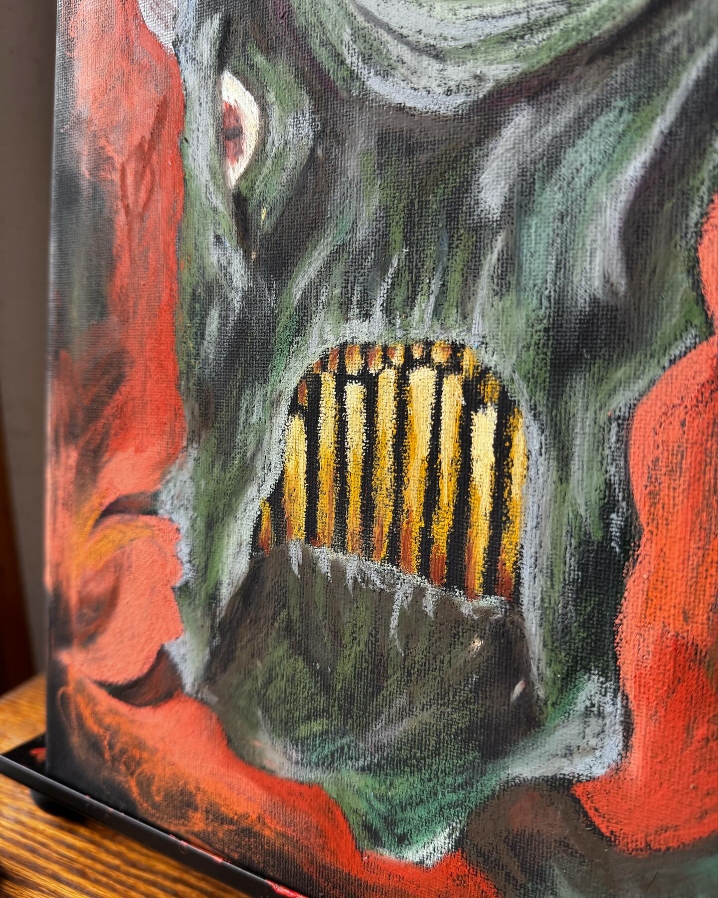 Close up photo of an art piece on an easel, green fantastical monster with horns, yellow teeth, and a very large eye. bckground is black and orange, almost entirely oil pastel.