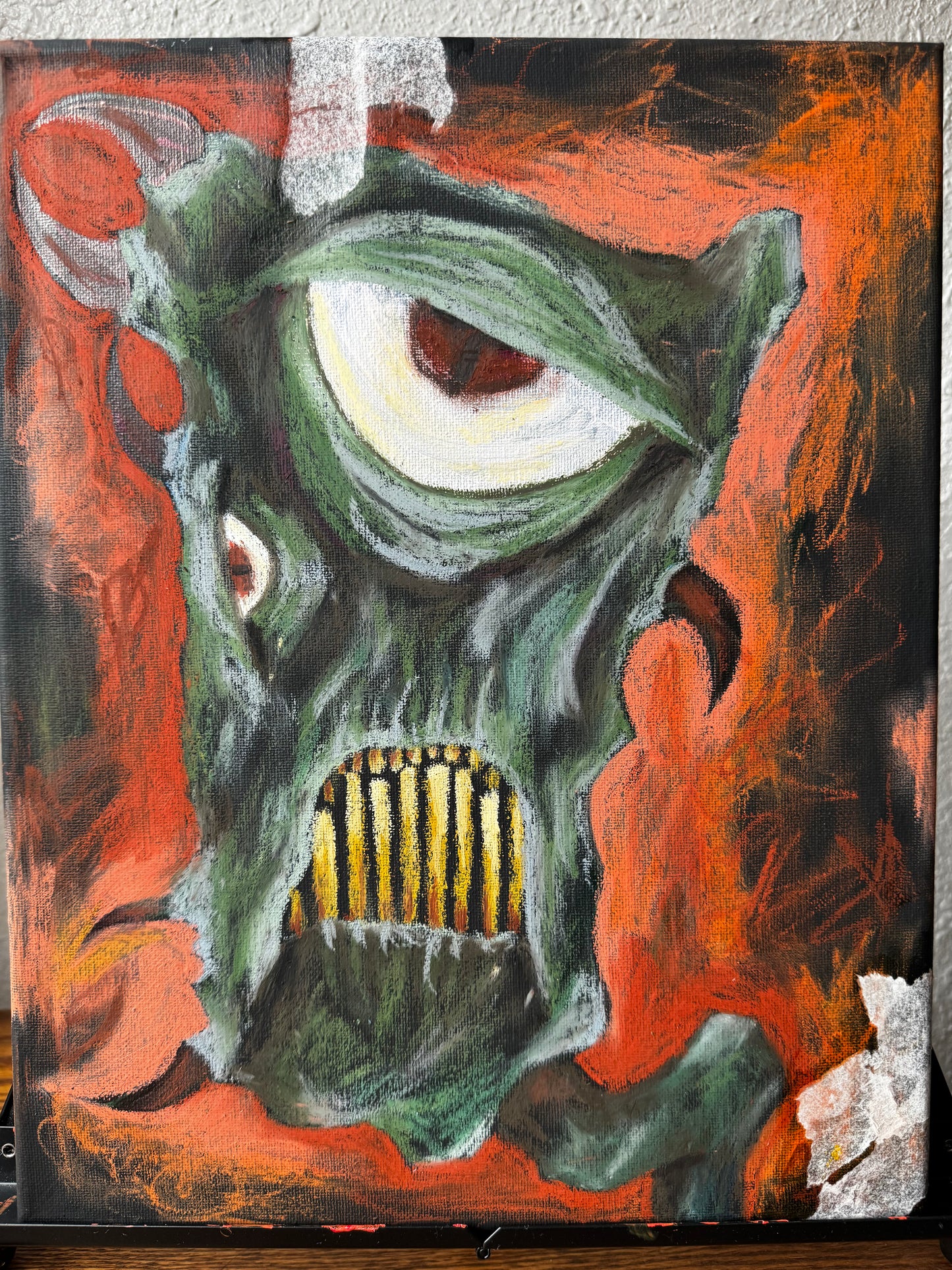 Straight on photo of an art piece on an easel, green fantastical monster with horns, yellow teeth, and a very large eye. bckground is black and orange, almost entirely oil pastel.