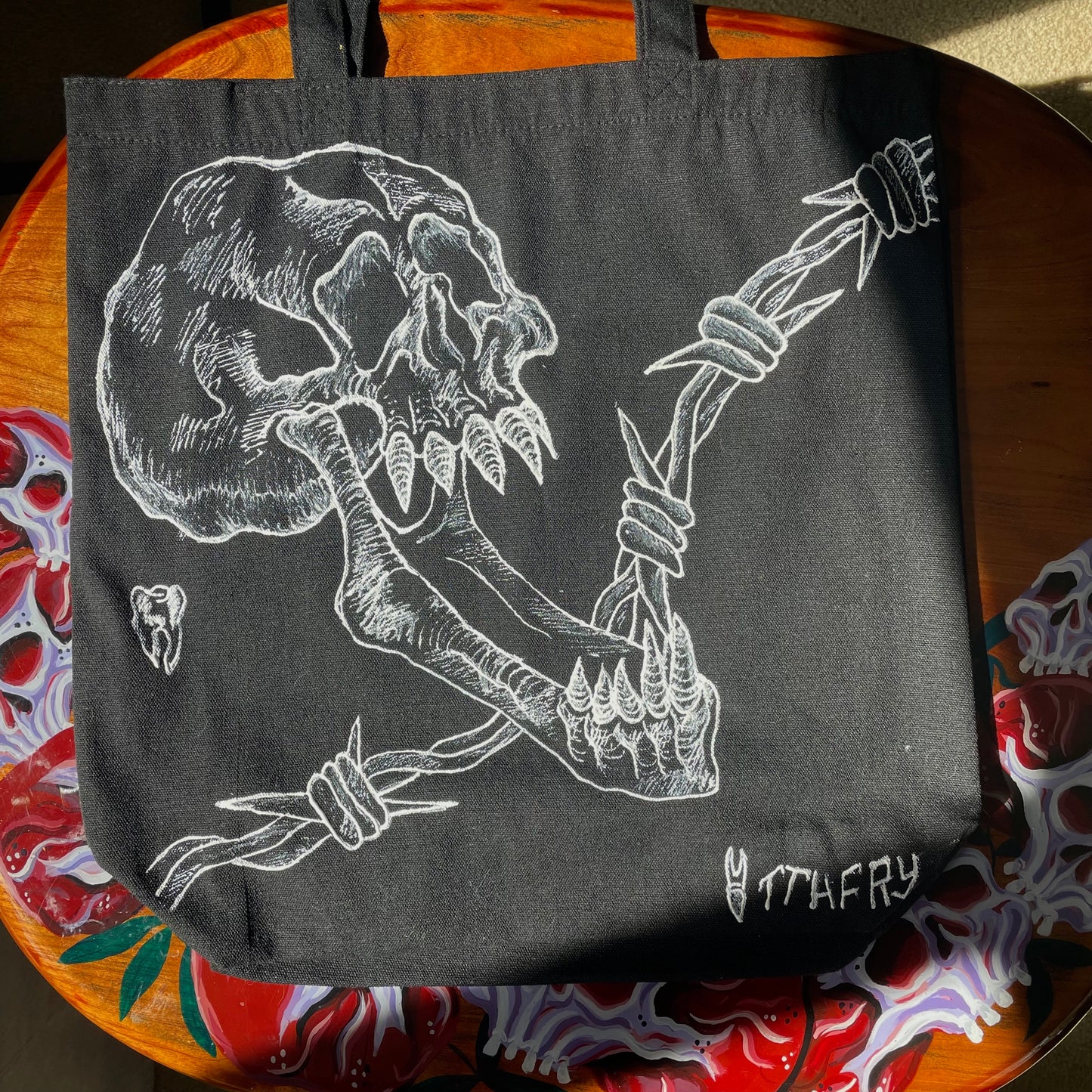 Straight on shot of black customized tote bag on a wooden painted skull table. Tote bag has a white fabric painted skull with a long jaw on it, barbed wire in the backgrund, and two teeth