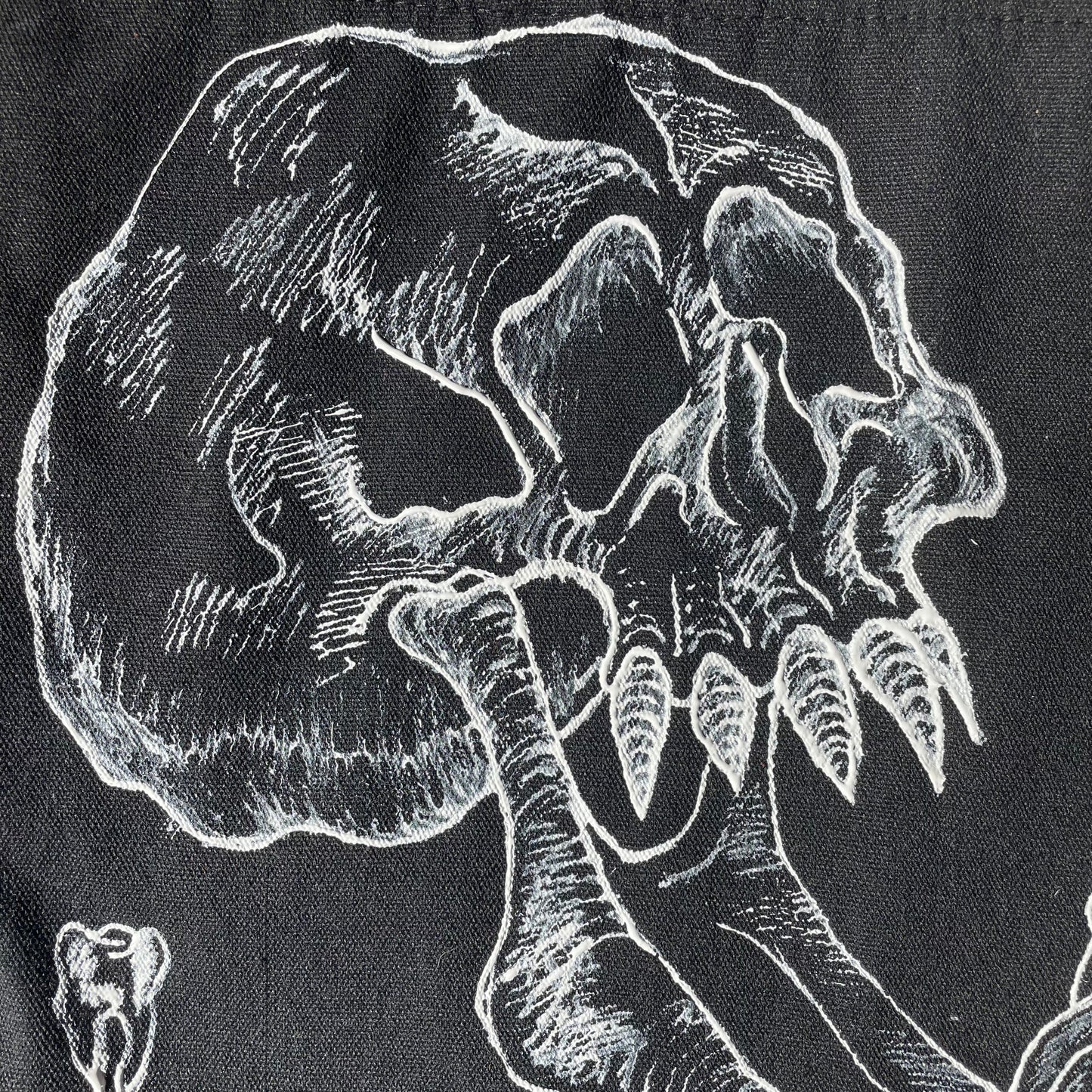 Close up shot of a black customized tote bag on a wooden painted skull table. Tote bag has a white fabric painted skull with a long jaw on it, barbed wire in the backgrund, and two teeth