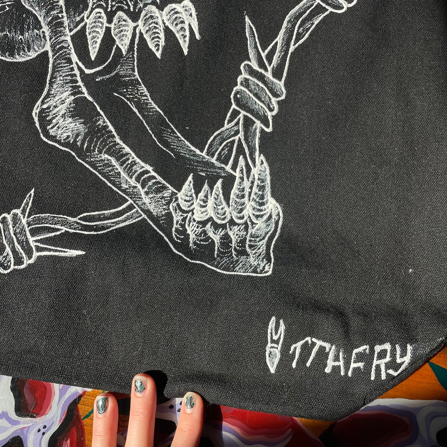 Another detail photo of a black customized tote bag on a wooden painted skull table. Tote bag has a white fabric painted skull with a long jaw on it
