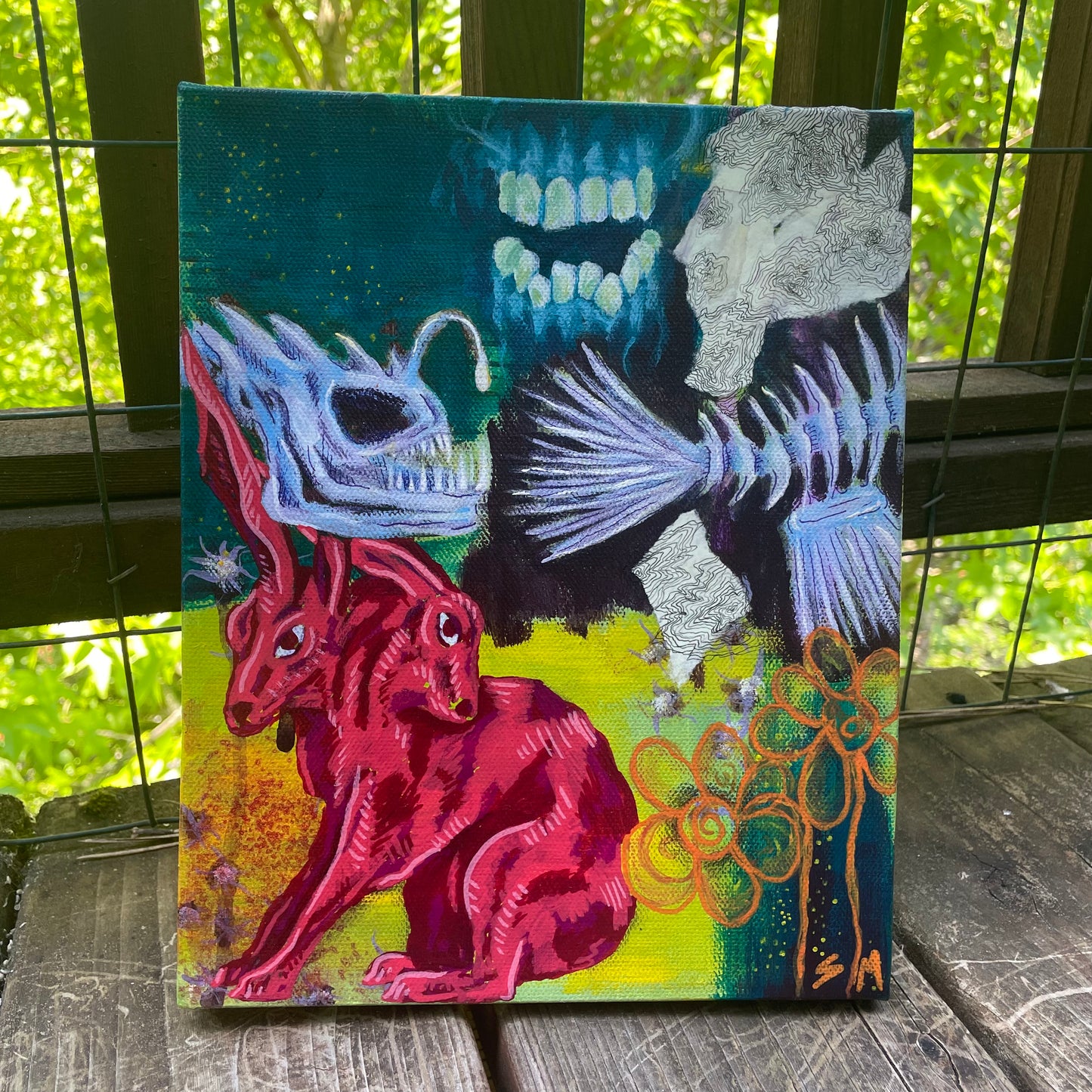 Photo of a painting sitting on a deck. Whimsical, original mixed-media painting, trippy two-headed rabbit artwork, angler fish skeleton, teeth and gums, orange flowers.