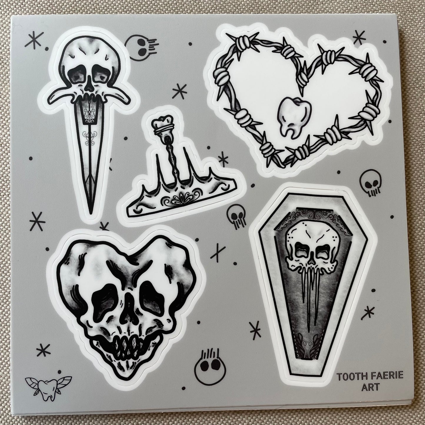 A sticker sheet of tattoo flash style drawings featuring a heart-shaped skull, a skull dagger, a tooth crown, a skull coffin, and a barbed wire heart with a tooth in the middle.