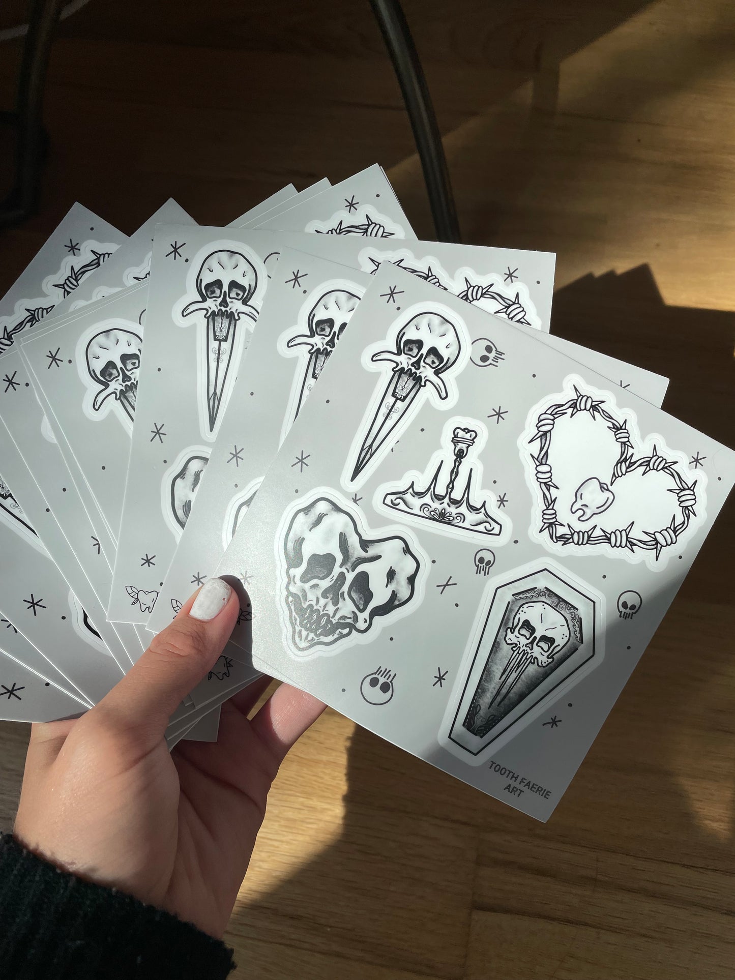 A hand holding a few sticker sheets of tattoo flash style drawings featuring a heart-shaped skull, a skull dagger, a tooth crown, a skull coffin, and a barbed wire heart with a tooth in the middle.