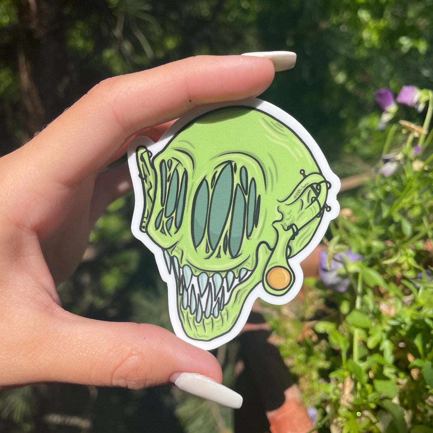 Green whimsical monster head sticker, cartoon style art, mythical creature with gauges and piercings. Hand holding up sticker against a garden background.