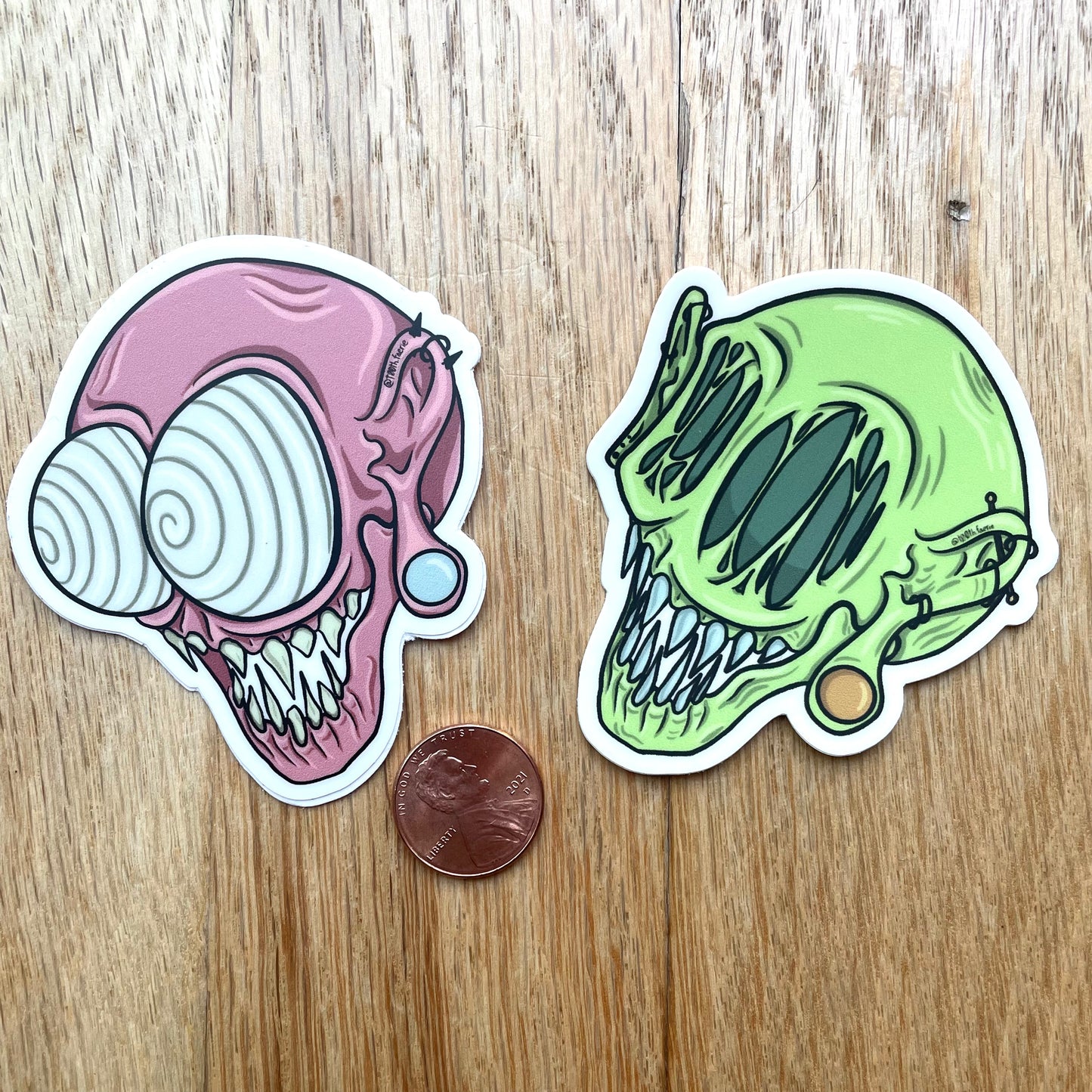 Two whimsical monster head stickers, cartoon style art, mythical creature with gauges and piercings. One green and one pink, cosmo and wanda style. on a table next to a penny