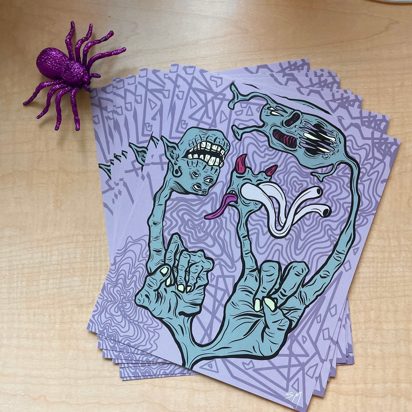 Poster prints spread out on a table next to a purple glittery spider. Prints are graphic, digital art style, showing two green hands. one hand holds up a pointer finger, which turns into a monster head. The hand on the right holds up the rock & roll sign (horns) and those fingers turn into other monster heads.