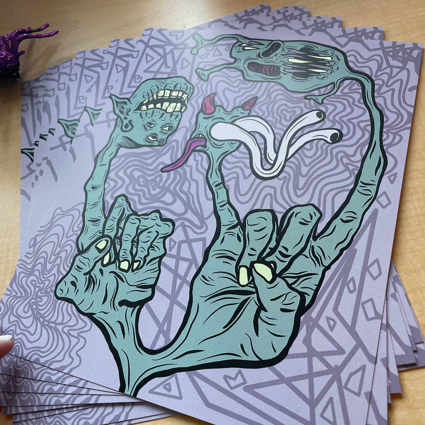 Angle shot of poster prints spread out on a table. Prints are graphic, digital art style, showing two green hands. one hand holds up a pointer finger, which turns into a monster head. The hand on the right holds up the rock & roll sign (horns) and those fingers turn into other monster heads.