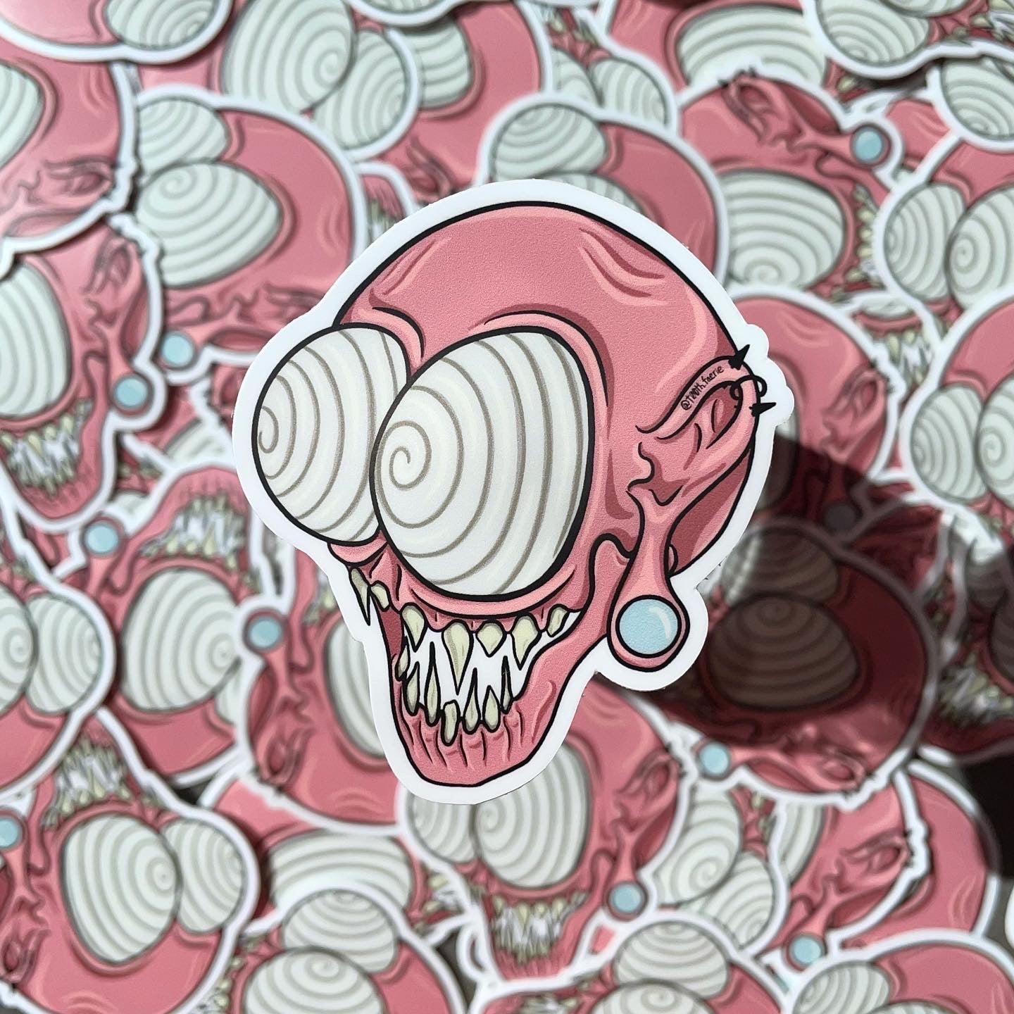Pink whimsical spooky monster head vinyl sticker, cartoon style art, mythical creature with gauges and piercings. One large sticker in focus over a pile of the same stickers.