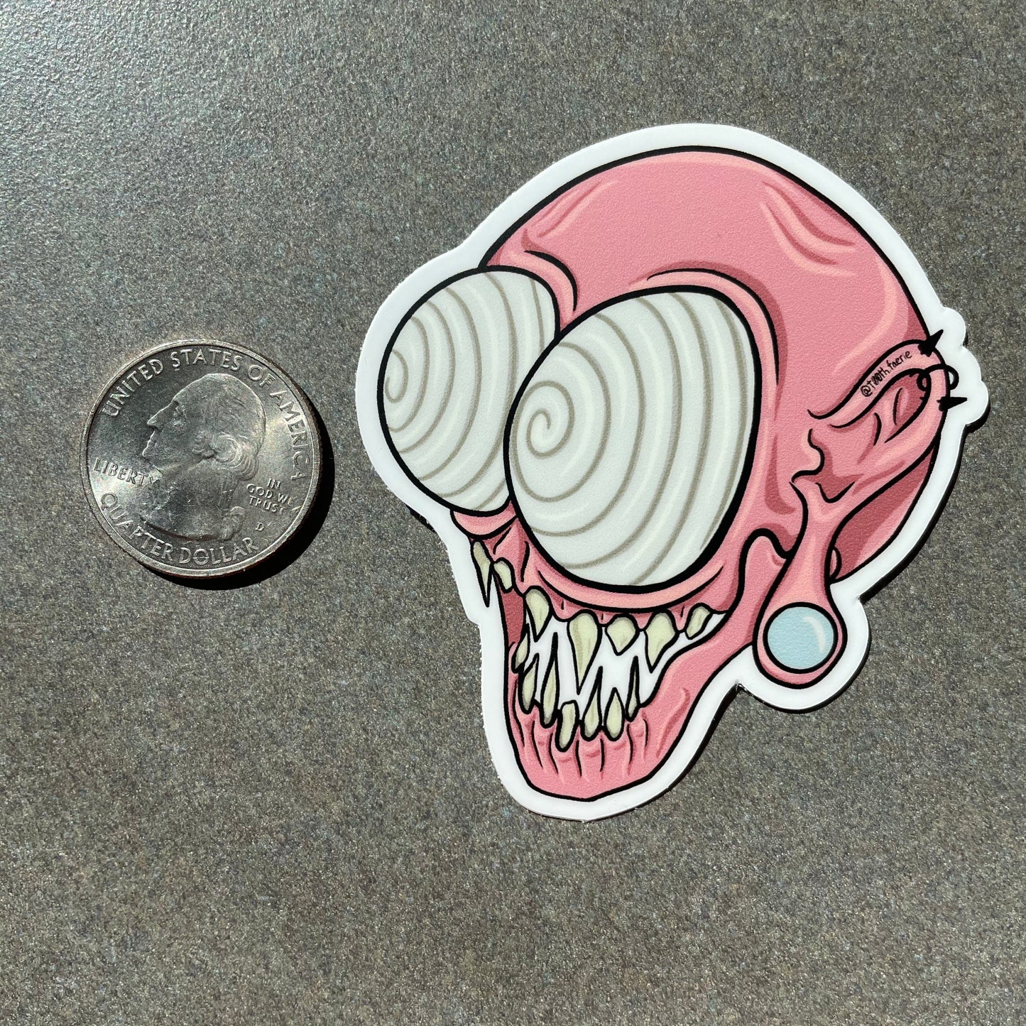 Pink whimsical spooky monster head vinyl sticker, cartoon style art, mythical creature with gauges and piercings. One sticker on a table next to a quarter for size comparison
