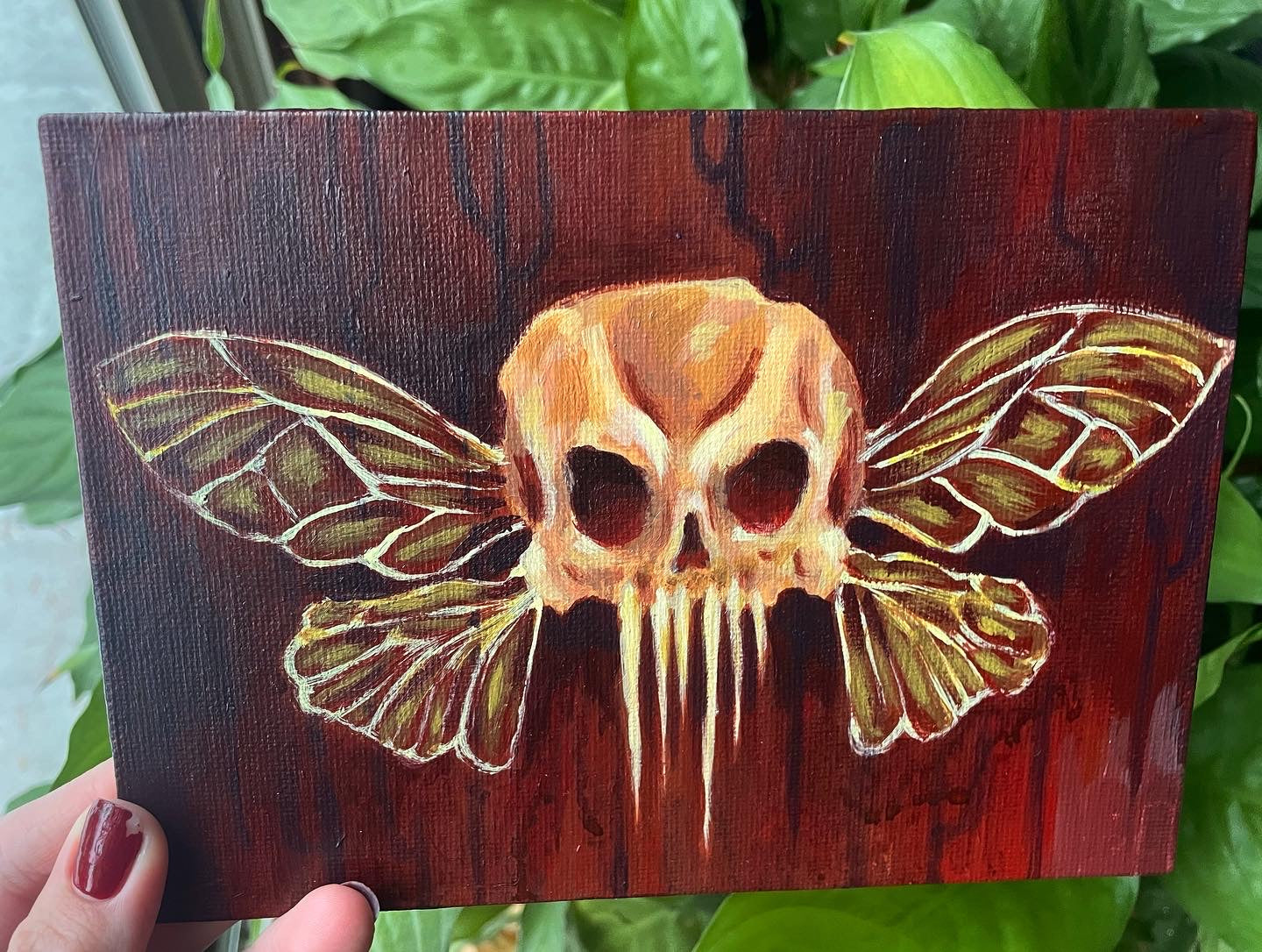 Hand holding up a small, spooky acrylic painting of a skull with fairy insect wings over a dark red, drippy background.