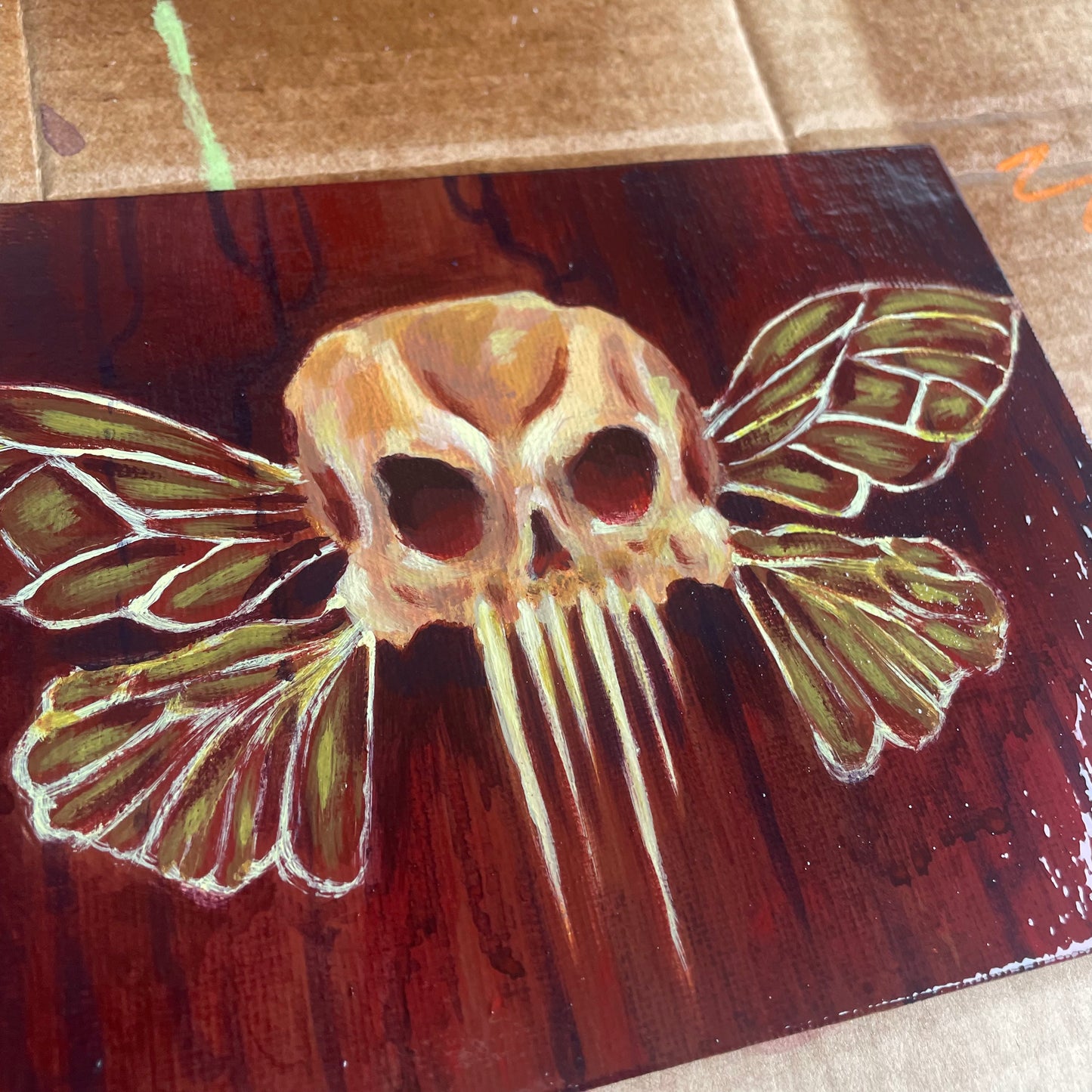 Close up of a small, spooky acrylic painting of a skull with fairy insect wings over a dark red, drippy background.