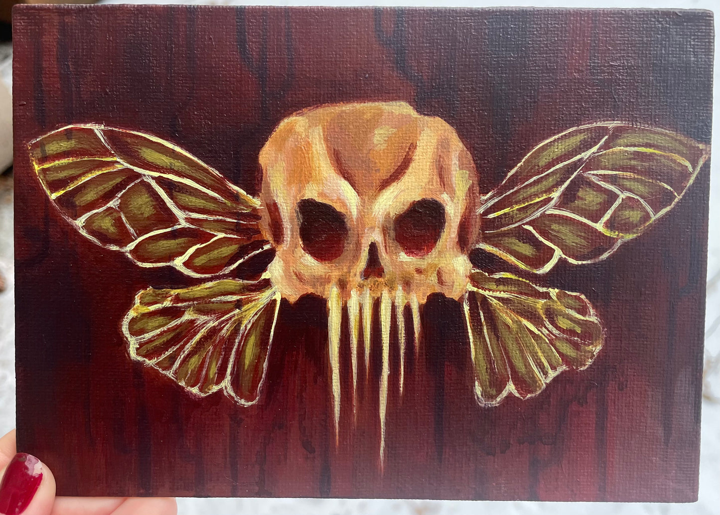Second angle, hand holding up a small, spooky acrylic painting of a skull with fairy insect wings over a dark red, drippy background.