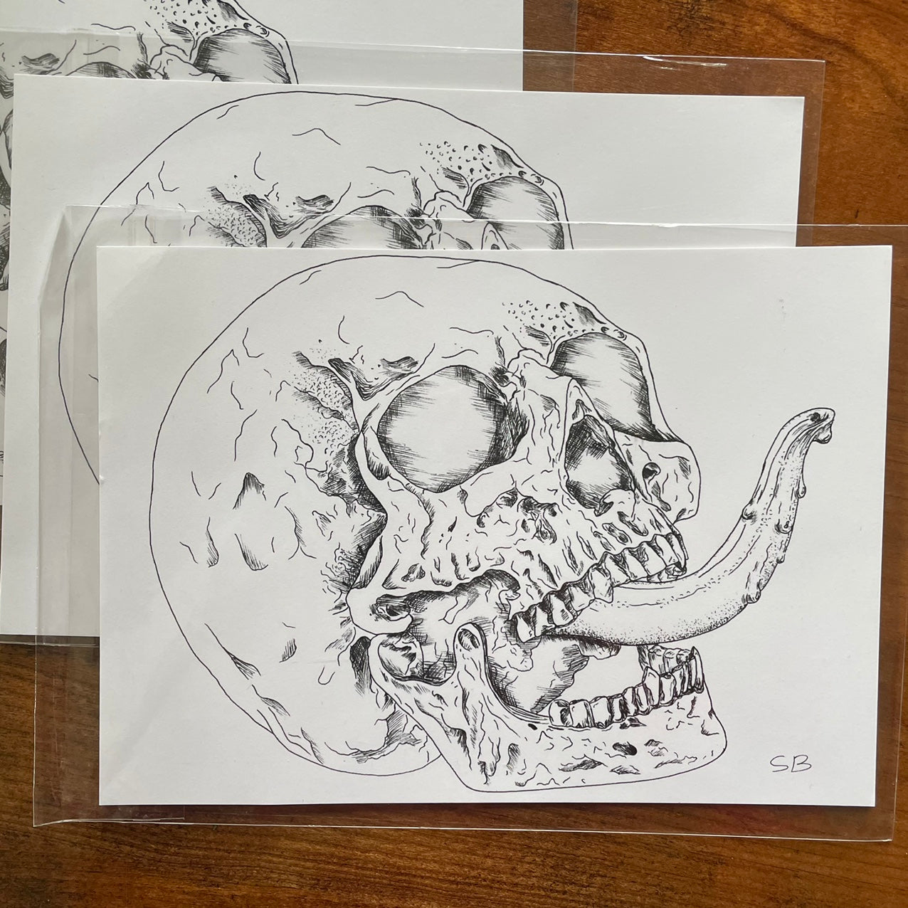 Photo of art prints on a table. Print depicts a drawing of a skull from the 3/4 (three quarter) angle with a tongue sticking out.
