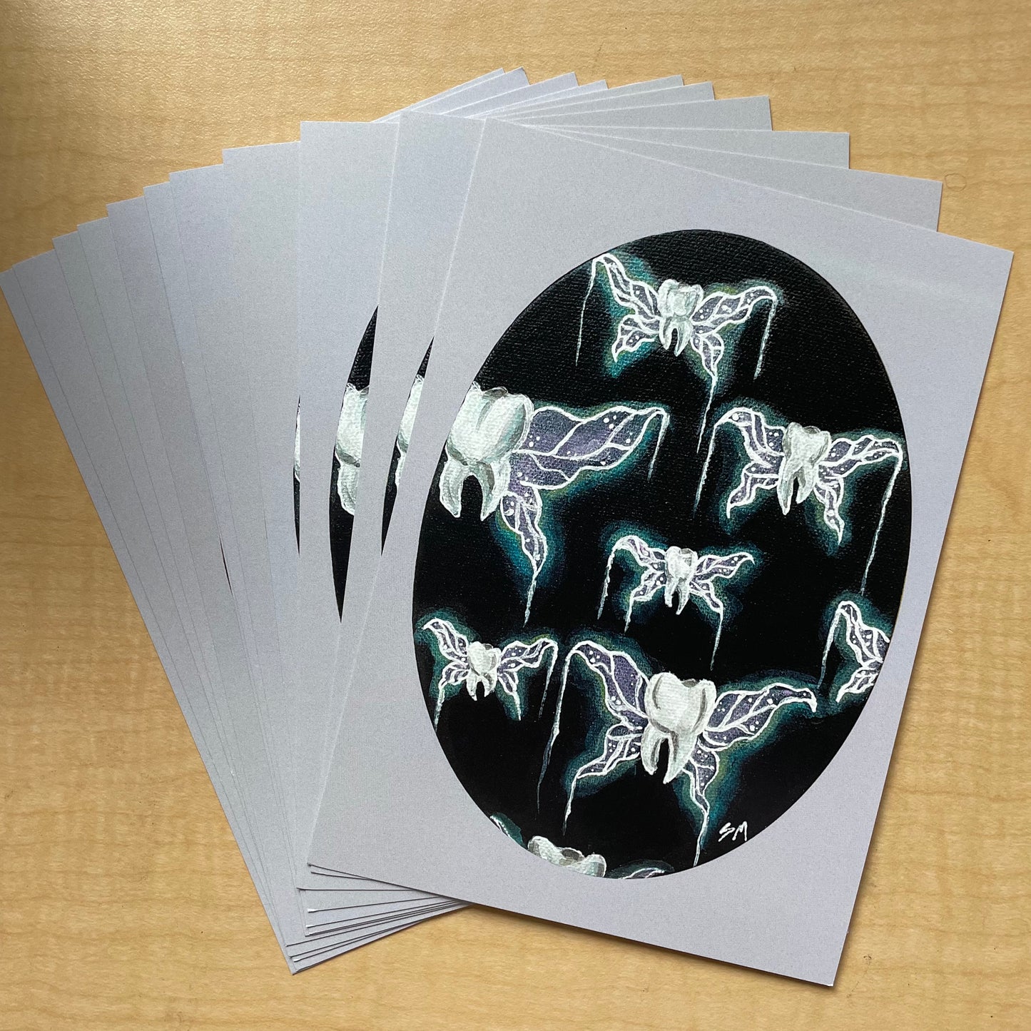 A spread of "tooth fairies" acrylic painting prints - teeth with glowing purple and teal fairy or insect wings suspended against a black background. Painting is an oval shape with a lavender border.