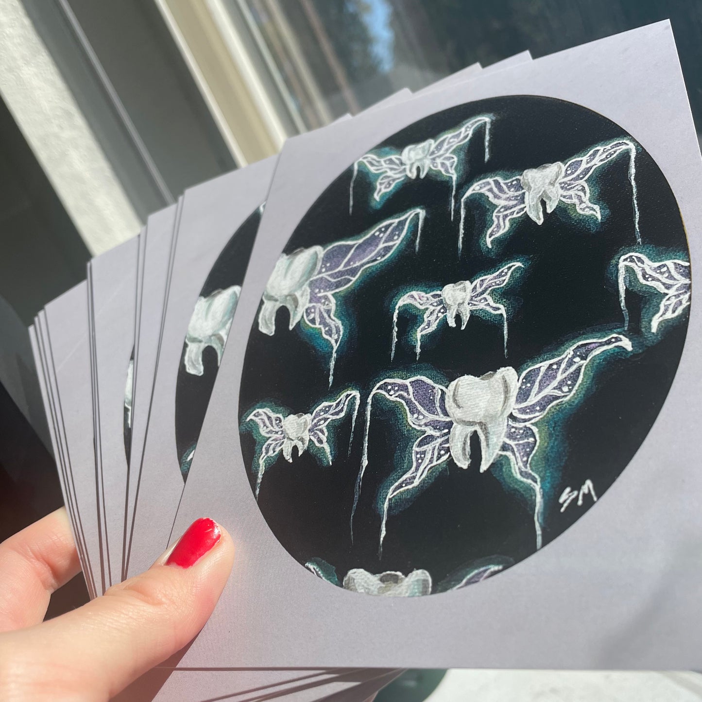 Hand holding a spread of prints of "tooth fairies" acrylic painting - teeth with glowing purple and teal fairy or insect wings suspended against a black background. Painting is an oval shape with a lavender border.