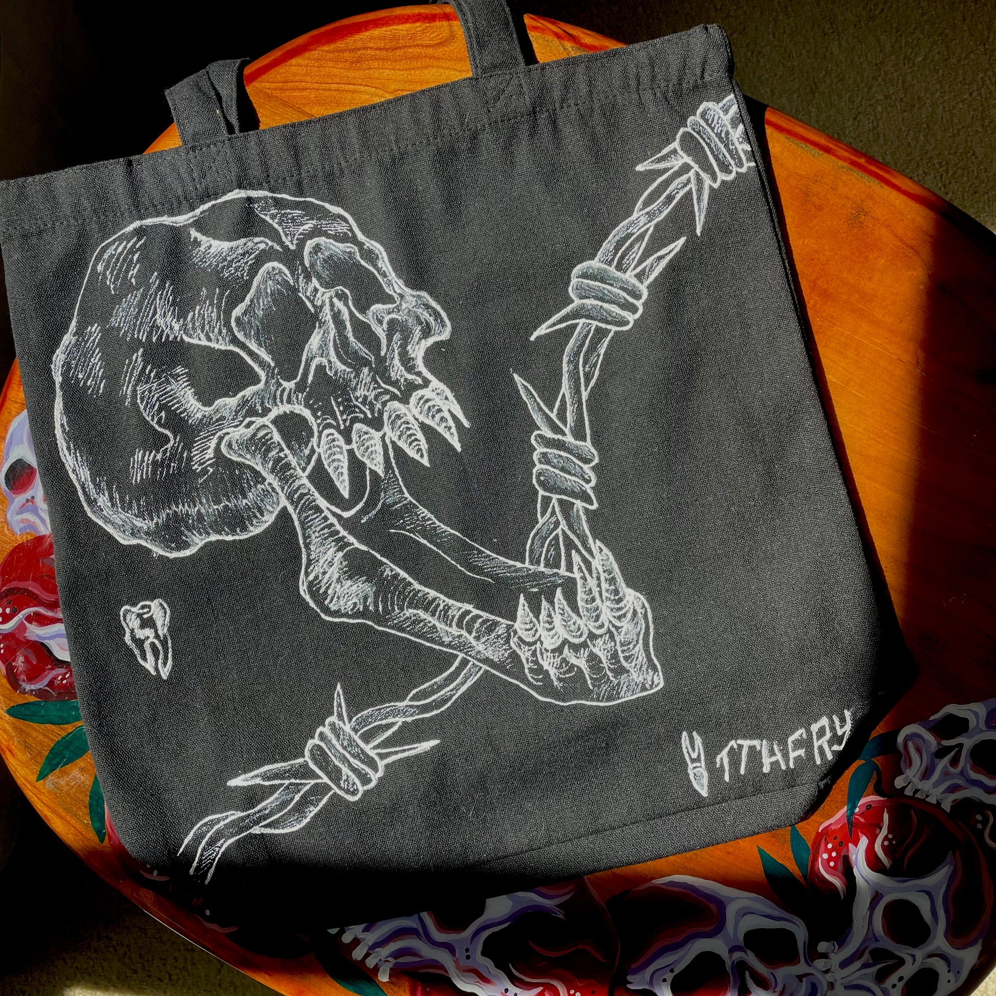 Black customized tote bag on a wooden painted skull table. Tote bag has a white fabric painted skull with a long jaw on it, barbed wire in the backgrund, and two teeth 