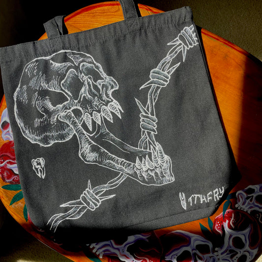 Black customized tote bag on a wooden painted skull table. Tote bag has a white fabric painted skull with a long jaw on it, barbed wire in the backgrund, and two teeth 
