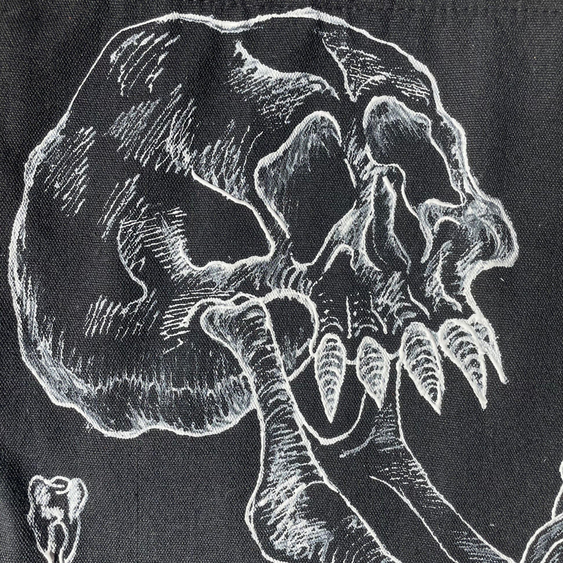 Close up shot of a black customized tote bag on a wooden painted skull table. Tote bag has a white fabric painted skull with a long jaw on it, barbed wire in the backgrund, and two teeth 