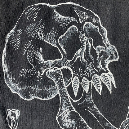 Close up shot of a black customized tote bag on a wooden painted skull table. Tote bag has a white fabric painted skull with a long jaw on it, barbed wire in the backgrund, and two teeth 