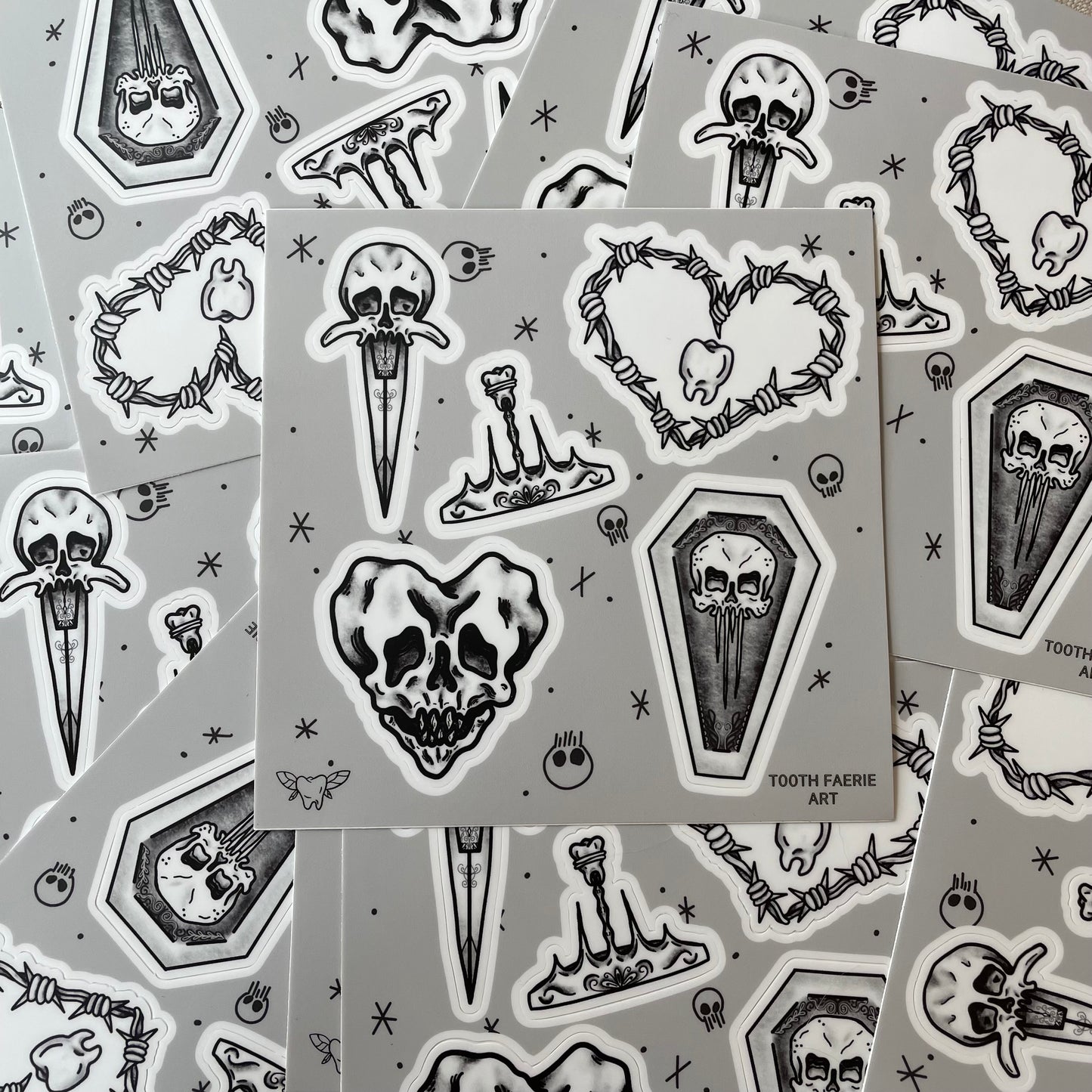 A pile of sticker sheets. Stickers are tattoo flash style featuring a heart-shaped skull, a skull dagger, a tooth crown, a skull coffin, and a barbed wire heart with a tooth in the middle.