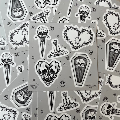 A pile of sticker sheets. Stickers are tattoo flash style featuring a heart-shaped skull, a skull dagger, a tooth crown, a skull coffin, and a barbed wire heart with a tooth in the middle.