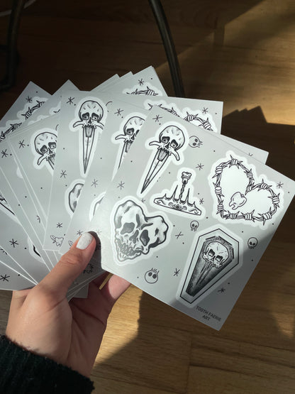 A hand holding a few sticker sheets of tattoo flash style drawings featuring a heart-shaped skull, a skull dagger, a tooth crown, a skull coffin, and a barbed wire heart with a tooth in the middle.
