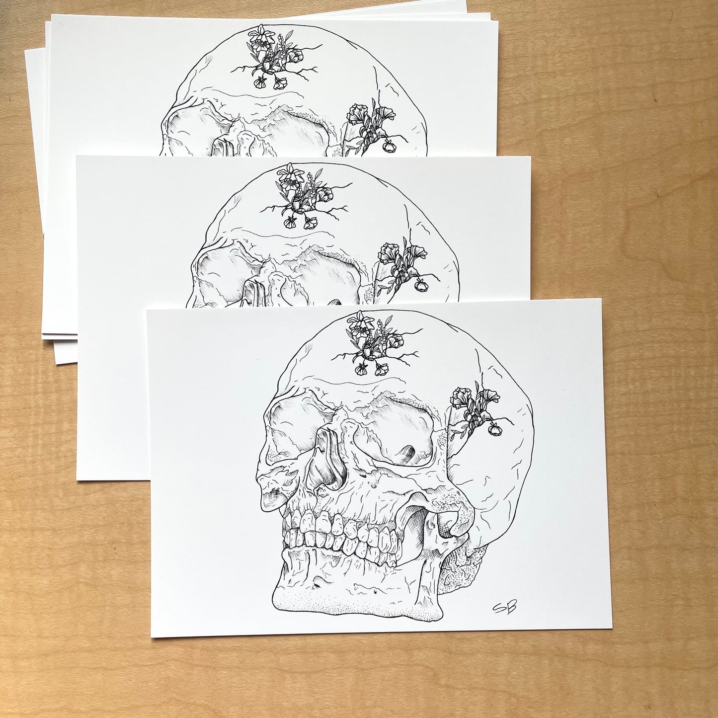 Multiple prints laying on a table of an ink drawing of a skull with cracks in it and flowers growing out of it.