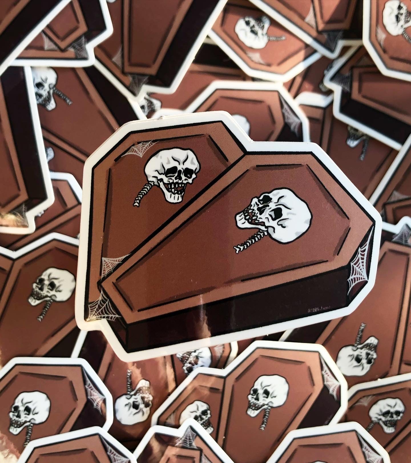 Photo of a vinyl sticker, two coffins in the shape of a heart with a skull on each and some spiderweb detailing