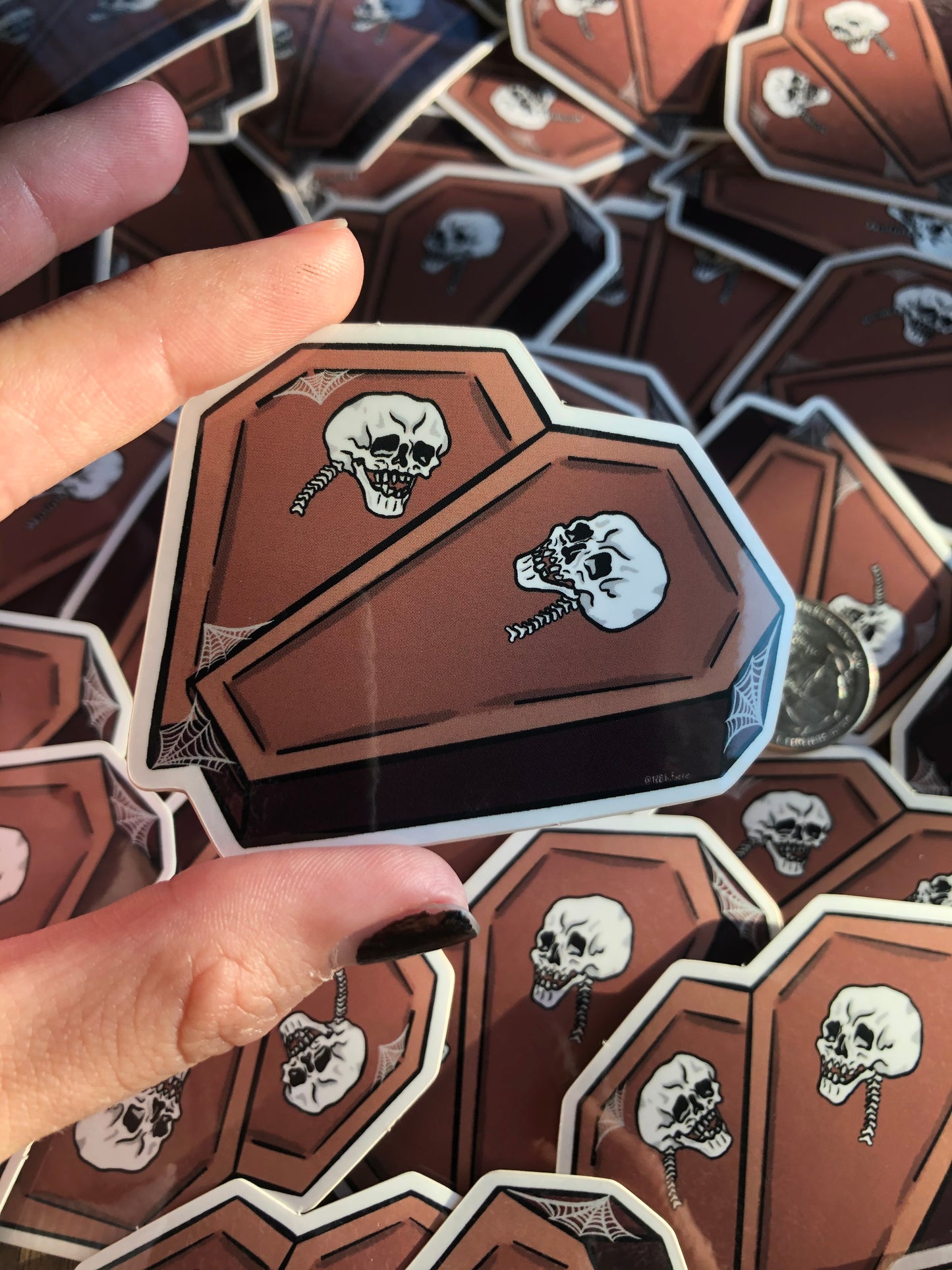 Hand holding a vinyl sticker depicting two coffins in the shape of a heart with a skull on each and some spiderweb detailing