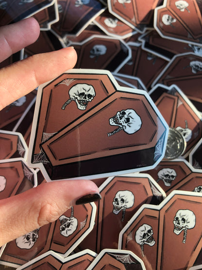 Hand holding a vinyl sticker depicting two coffins in the shape of a heart with a skull on each and some spiderweb detailing