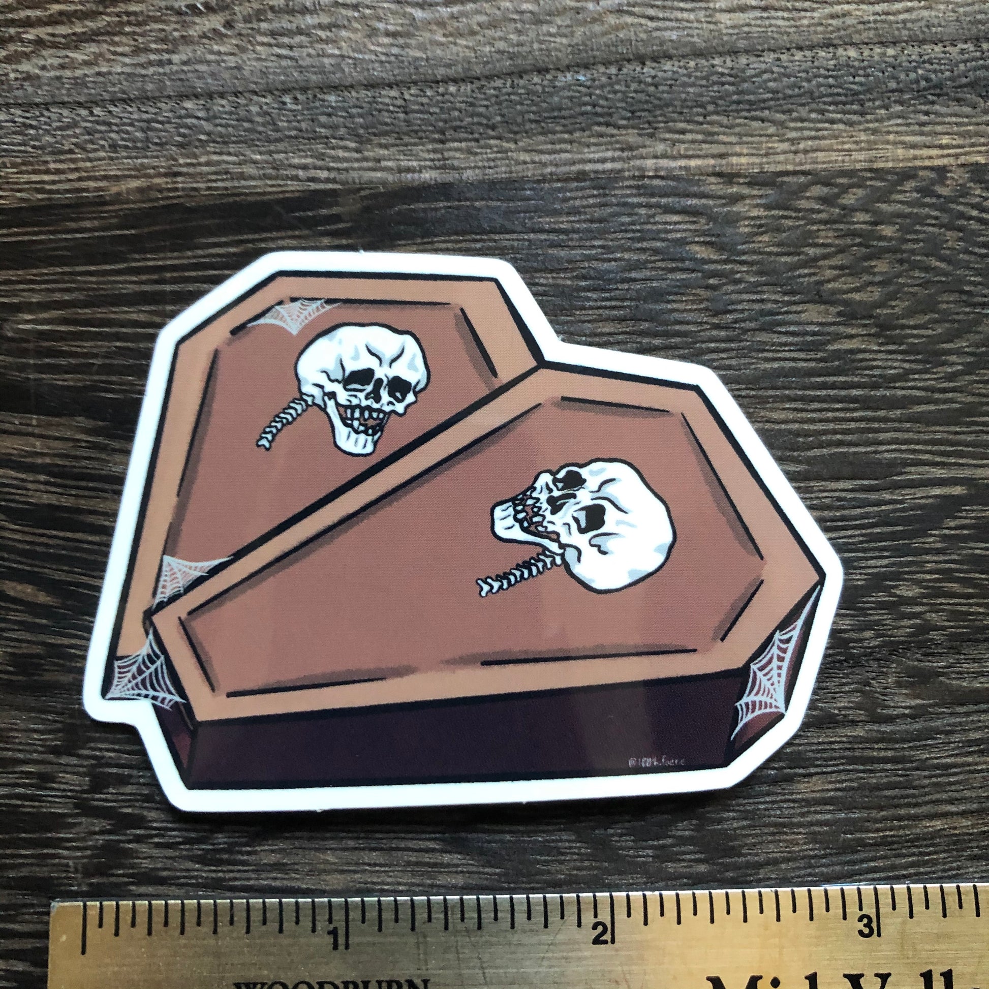 Photo of a vinyl sticker next to a ruler, sticker of two coffins in the shape of a heart with a skull on each and some spiderweb detailing