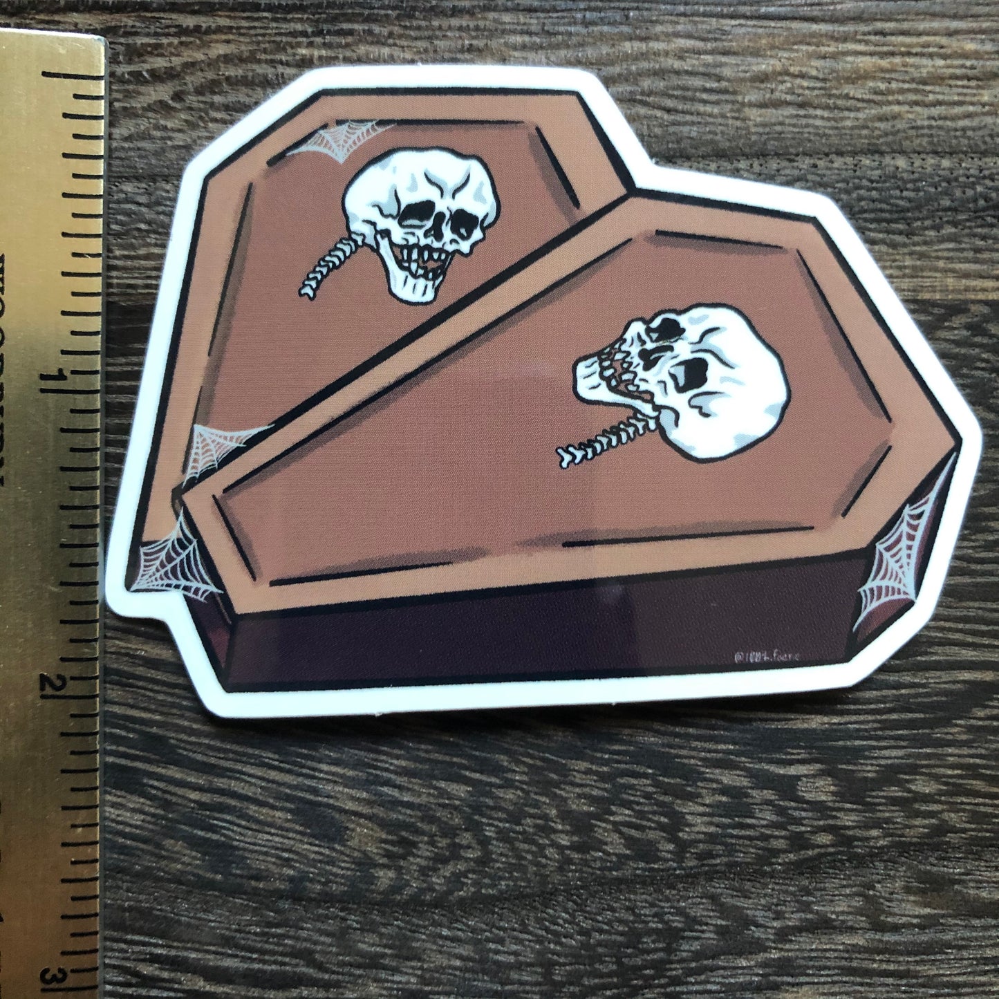 ruler showing height of a vinyl sticker of two coffins in the shape of a heart with a skull on each and some spiderweb detailing