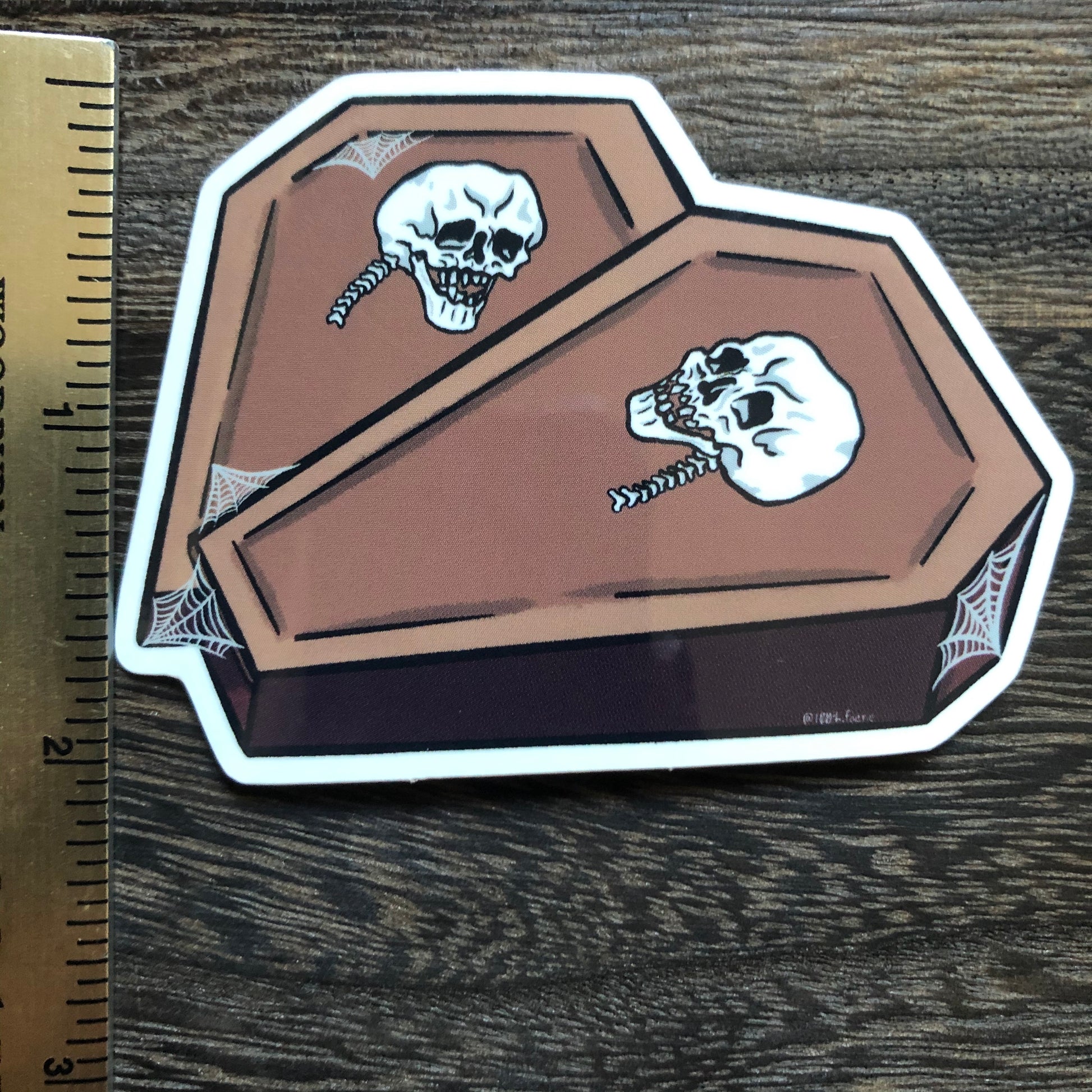 ruler showing height of a vinyl sticker of two coffins in the shape of a heart with a skull on each and some spiderweb detailing