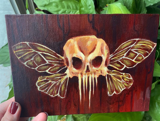 Hand holding up a small, spooky acrylic painting of a skull with fairy insect wings over a dark red, drippy background. 