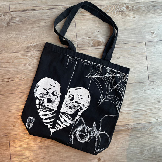 Black tote bag product photo adorned with two skulls in a heart, a spiderweb in the corner, a big spider, and a tooth in white fabric paint