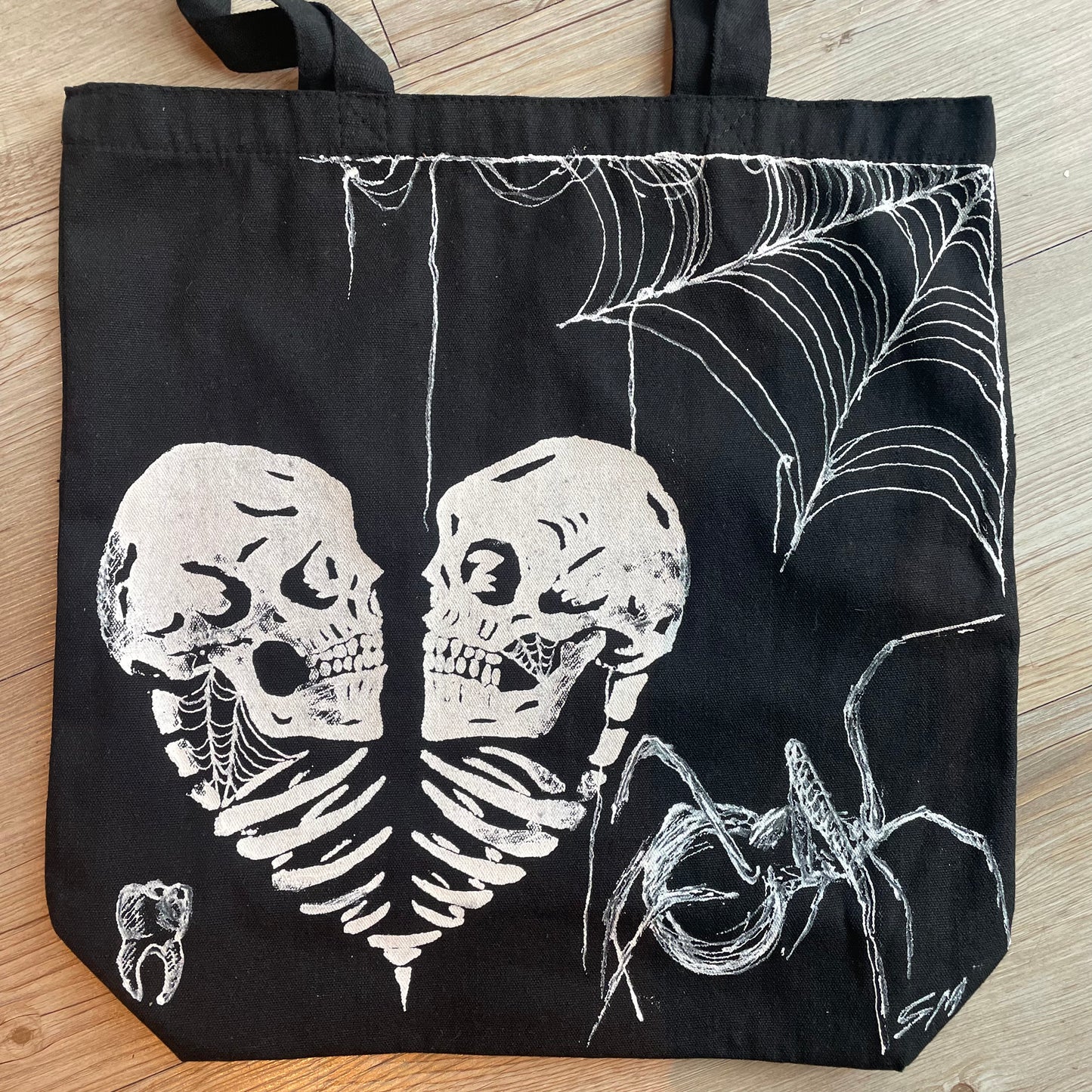 Straight on photo of a black tote bag product photo adorned with two skulls in a heart, a spiderweb in the corner, a big spider, and a tooth in white fabric paint