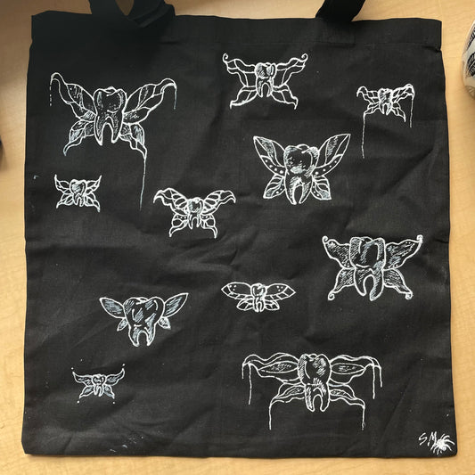 Black tote bag laying flat on table, customized with white fabric paint depicting "tooth fairies" - teeth with fairy wings.