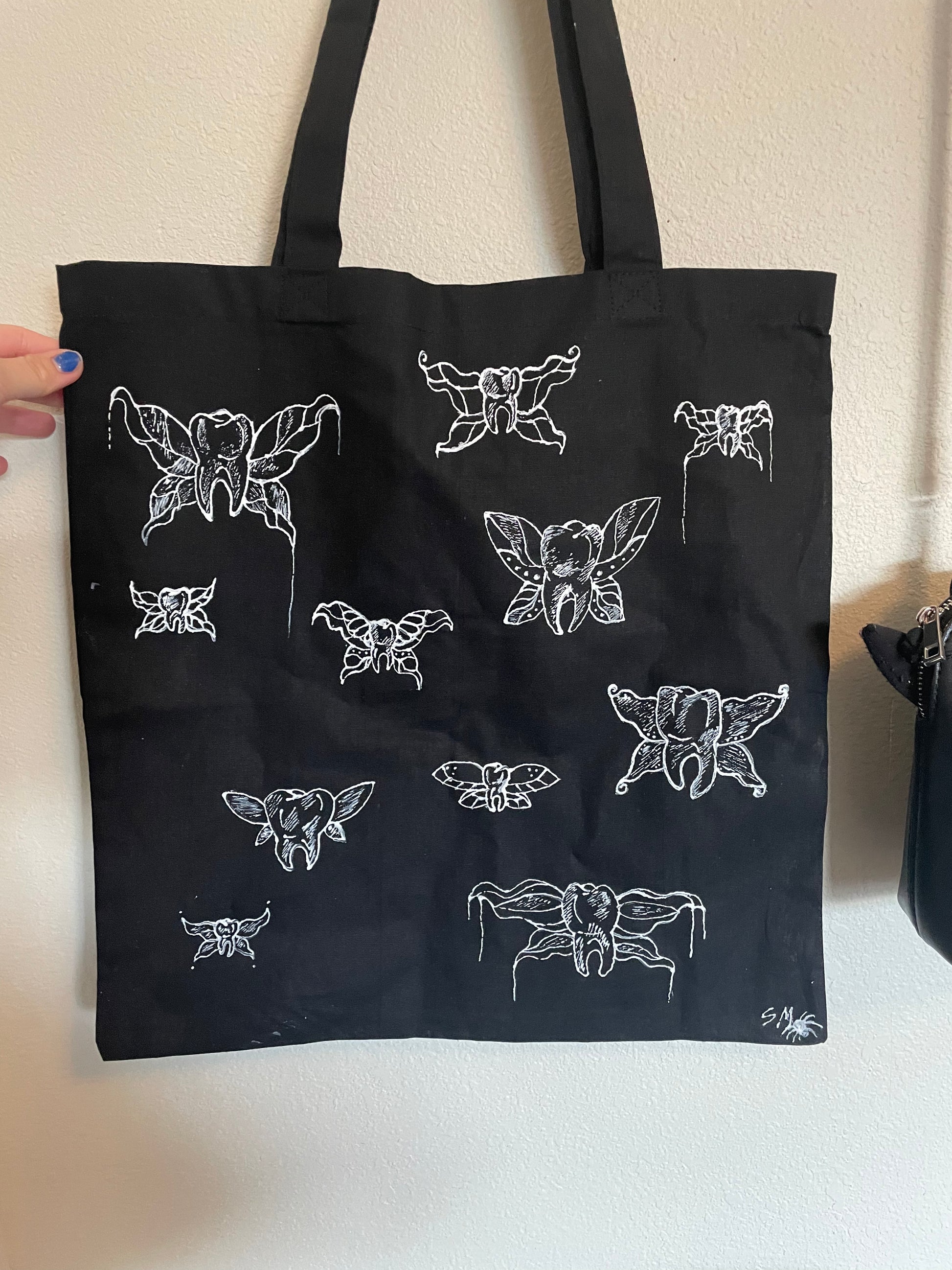 Black tote bag hanging against a white wall, customized with white fabric paint depicting "tooth fairies" - teeth with fairy wings.