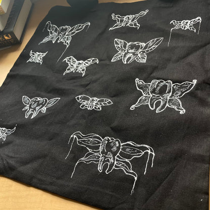 Close up show of black tote back laying flat on table, customized with white fabric paint depicting "tooth fairies" - teeth with fairy wings.