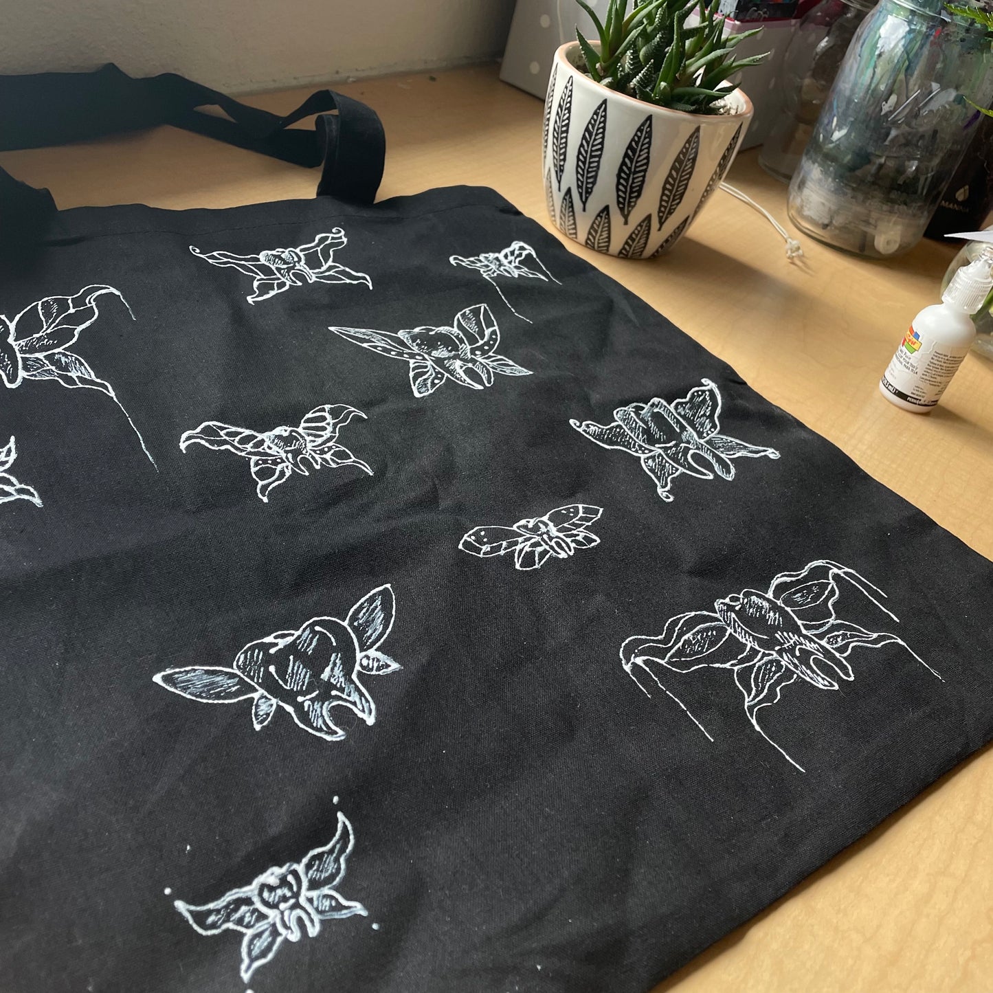 Side angle of black tote back laying flat on table, customized with white fabric paint depicting "tooth fairies" - teeth with fairy wings.